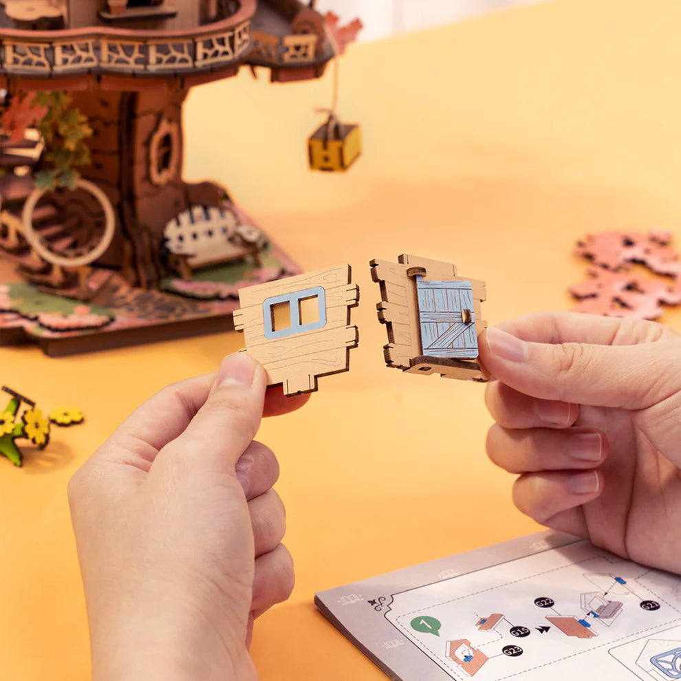 Two hands holding small wooden architectural models against a blurred background of a detailed wooden model set.